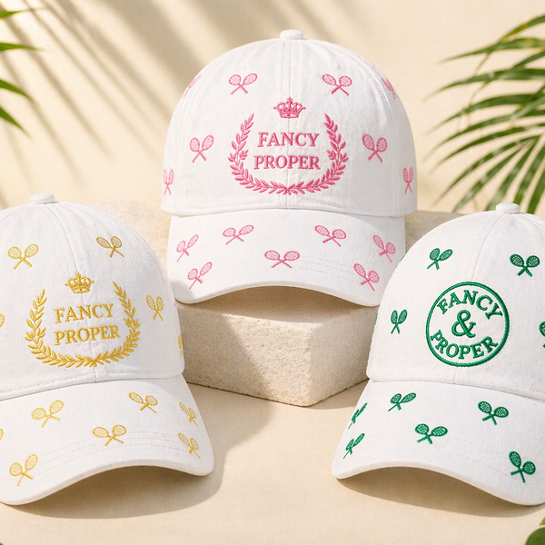 Three white baseball caps with embroidered designs on a beige surface with green leaves in the background.