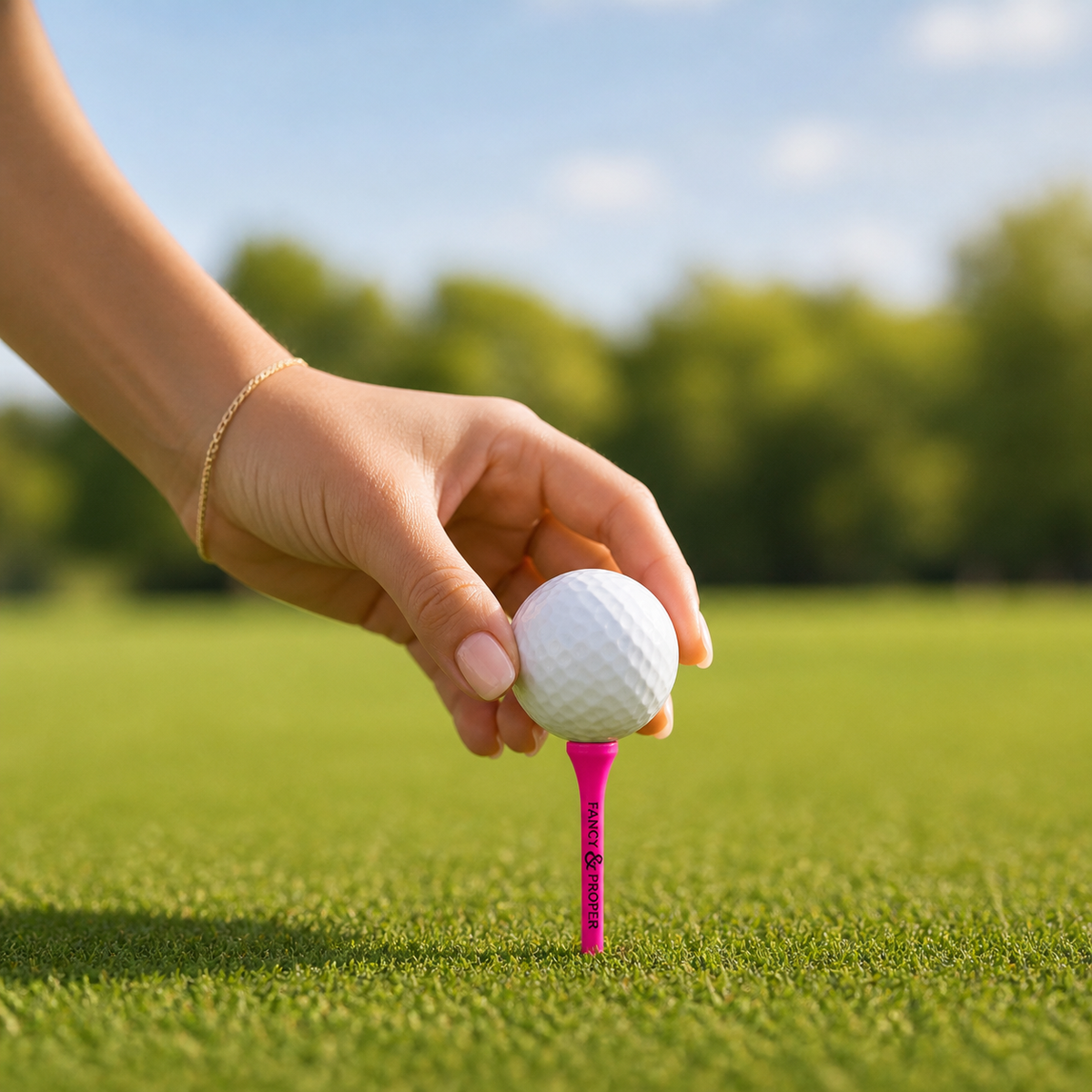 Hand placing a golf ball on a pink tee with a blurred green field and blue sky in the background
